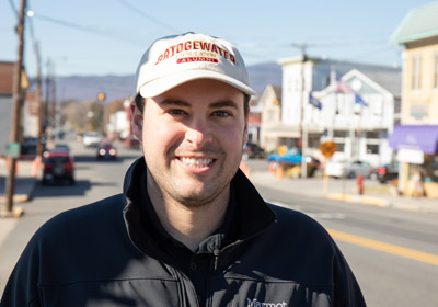 Man in foreground smiles at camera. Background is of a road and storefronts.|