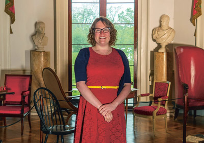 woman in red dress stands in the middle of a room|woman brushes artifacts on a display table|woman cleans a light fixture hanging from the ceiling|