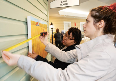 two young woman hang a painting with a tape measure||Young woman stands next to a painting of a human heart|Young woman sits behind pottery|Young woman hangs a painting on a wall with a tape measure|