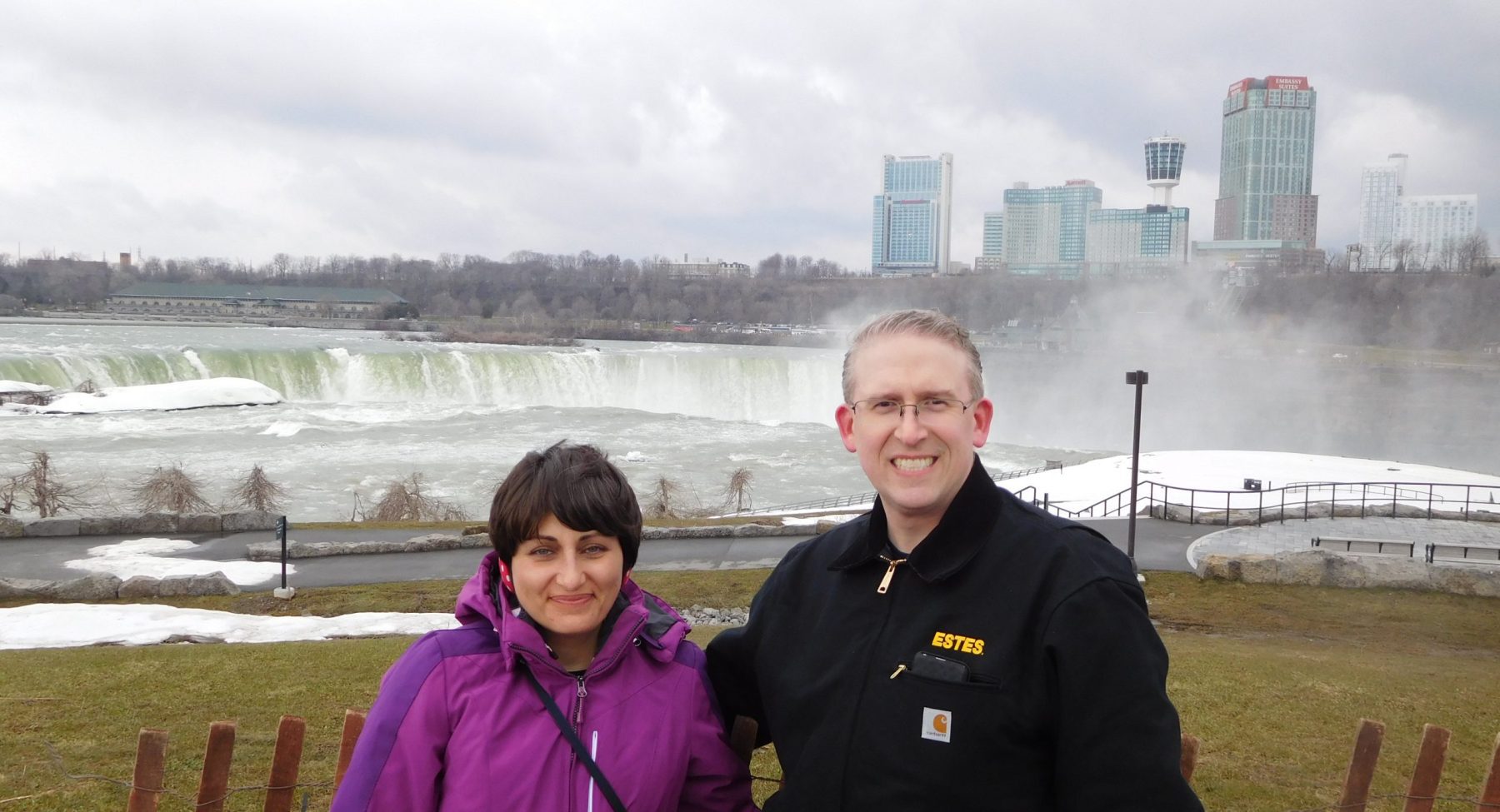 Woman and man stand in front of waterfall with skyline in background