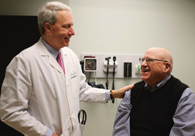 Doctor on left puts hand on shoulder of patient who is sitting on a table.|