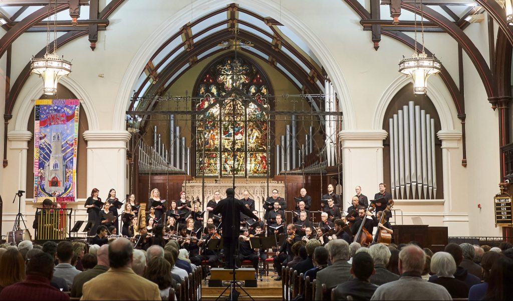 the back of a band director can be seen center stage with an orchesra before him. An audience sits in pews