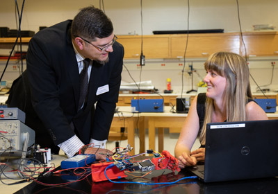 Man in suit stands stands next to student who is seated in front of a computer. A red robtics arm rests on the table between them