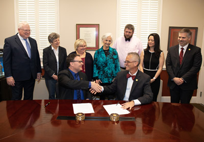 Two men sit at a desk with papers in front of them and shake hands while people stand behind them||