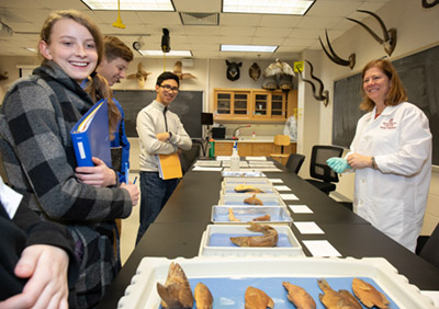 Three students stand on the left in front of boxes of fish specimen while Dr. Kimberly Bolyard stands on the right||Four high school students stand on the left while Dr. Tamara Johnstone-Yellin holds a skull.