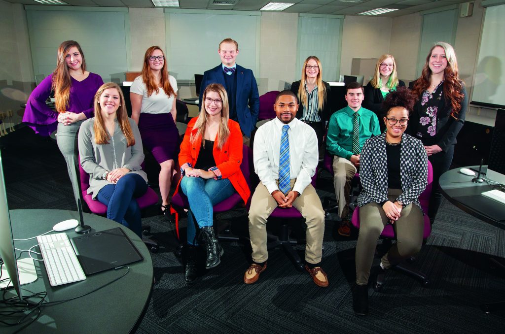 Five students sit in a computer lab while six students stand behind them