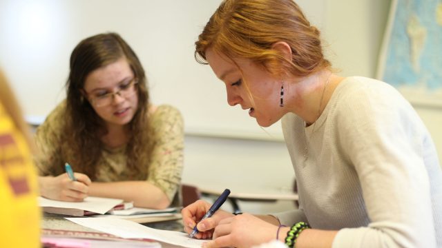 Female student writing on paper with another female student in the background