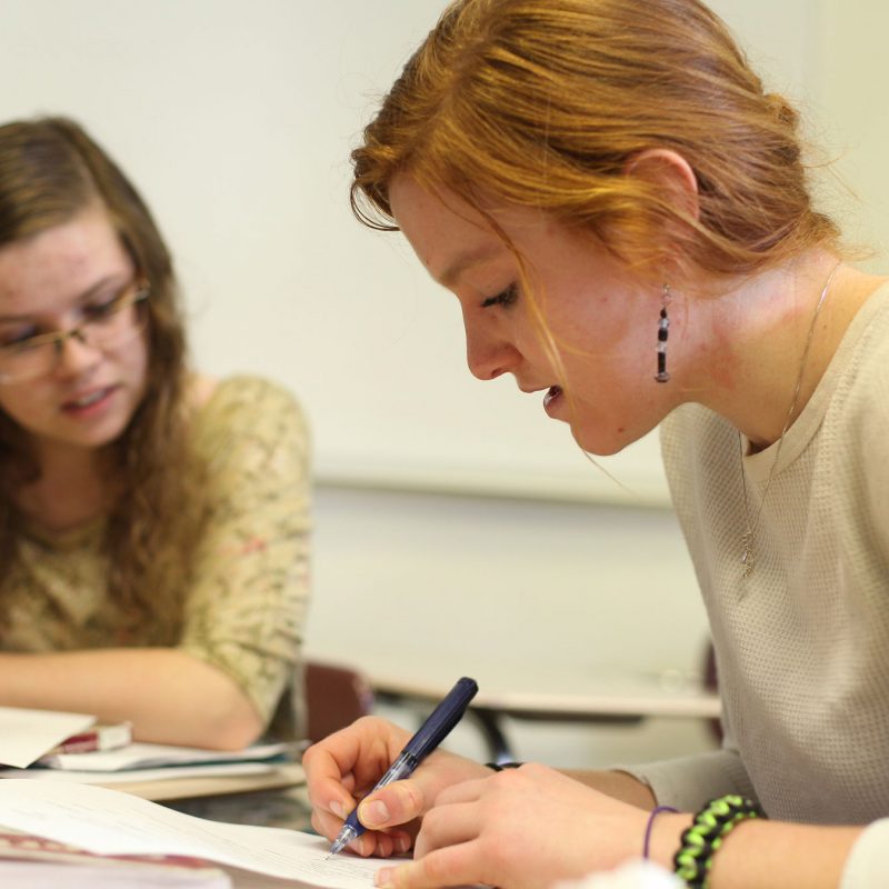 Female student writing on paper with another female student in the background