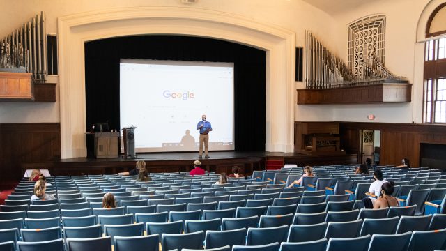 Man standing on stage with Google projected on a screen in an auditorium with a small amount of students