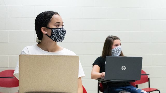 Two female students sitting at desks with laptops in front of them with masks covering their faces
