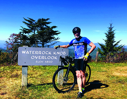 Man on bicycle stands next to sign that says Waterrock Knob Overlook Elev 5820