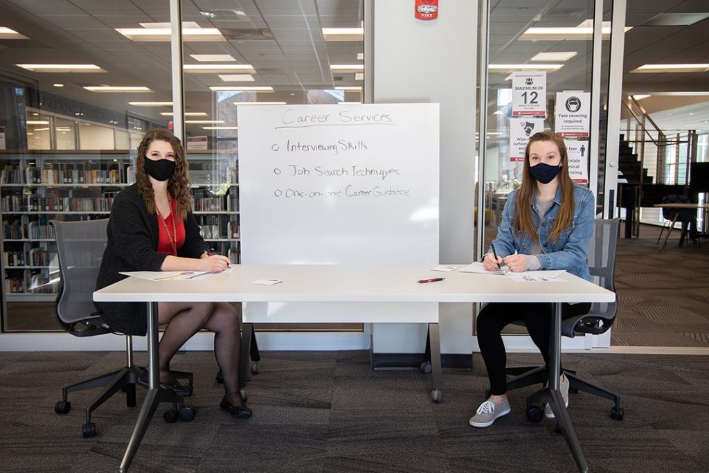 Two females sitting at a desk with white board in the middle with text about career services