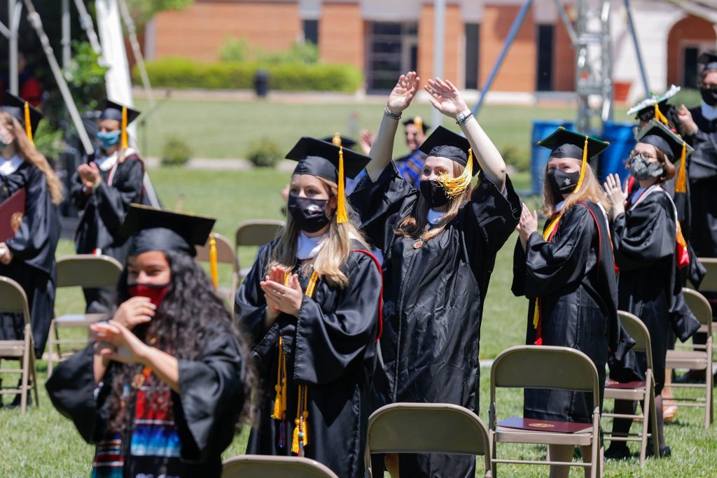 Students wearing graduation robes stand by their chairs and clap.
