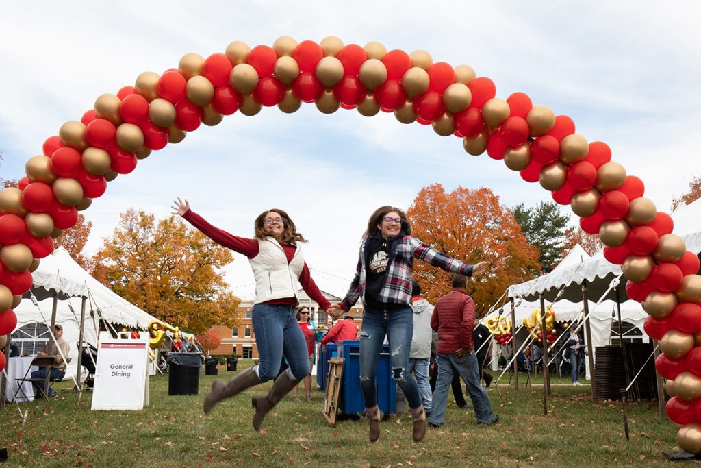Two girls in mid-air under gold and crimson balloon banner at Homecoming