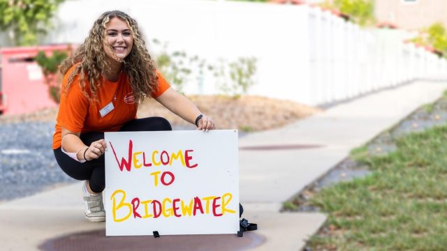A girl on the sidewalk holds a sign that reads Welcome To Bridgewater in red and gold writing