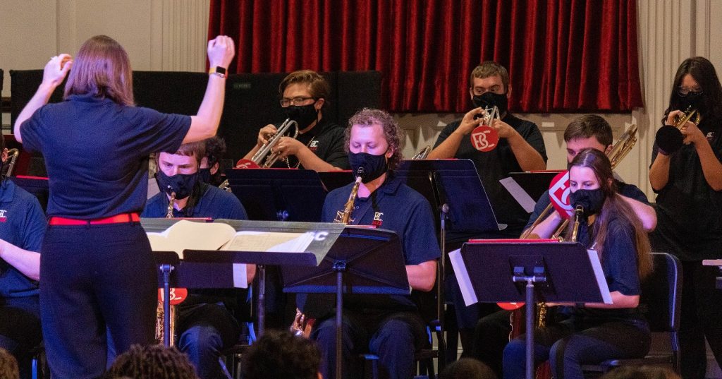 The back of a band director can be seen conducting a band of students. Performers are wearing all back and masks.