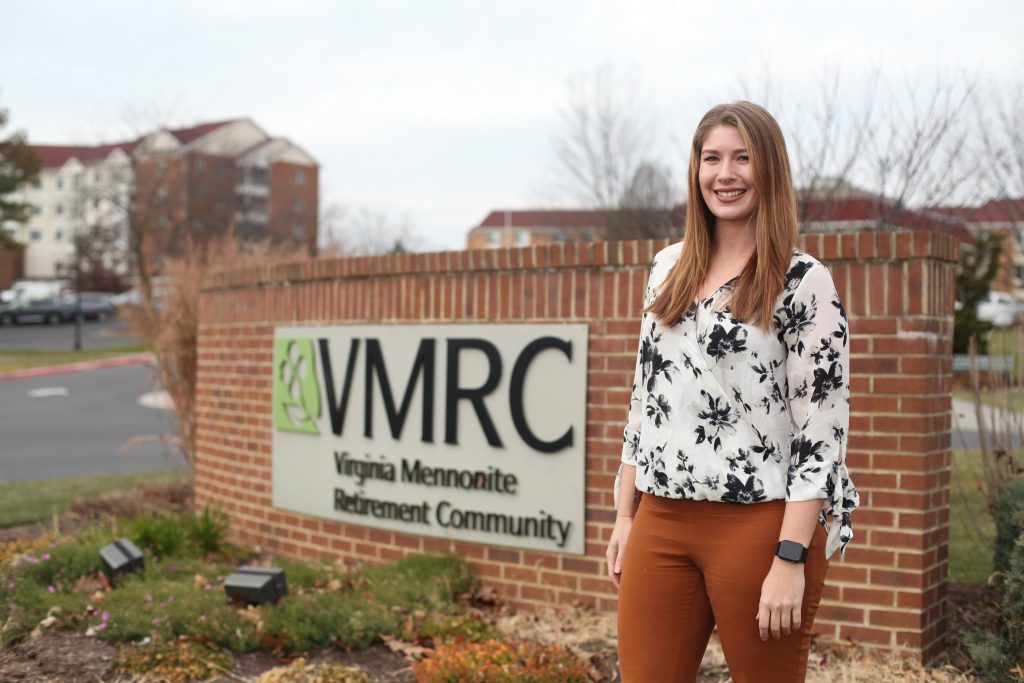 A woman stands in front of a sign that says V-M-R-C Virginia Mennonite Retirement Community