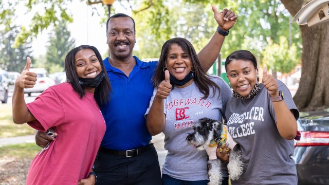 A family of four and their dog pose for a photo smiling and with their thumbs up