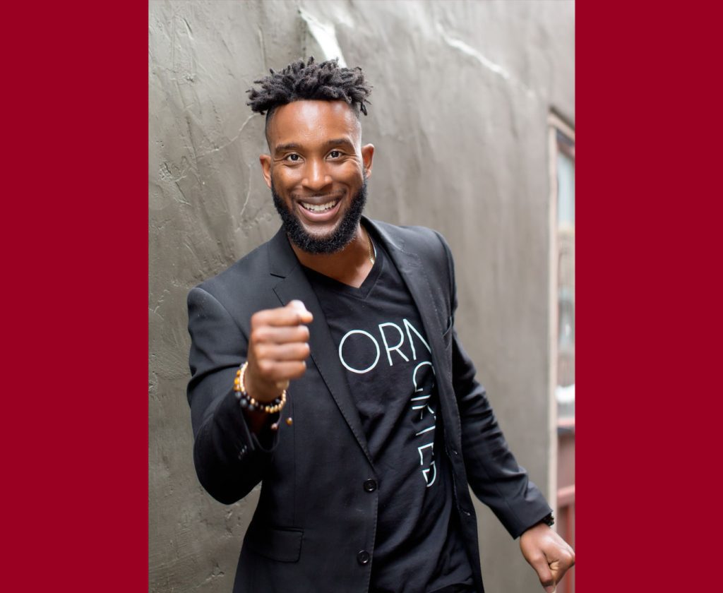 A man in a black suit stands against a gray wall with his hand up in a fist and smiling