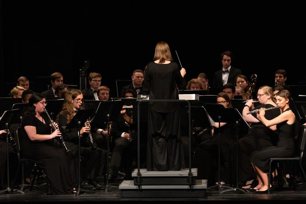 A band director is center stage with her back to the audience. Band members playing various woodwind instruments face her. They are all dressed in black