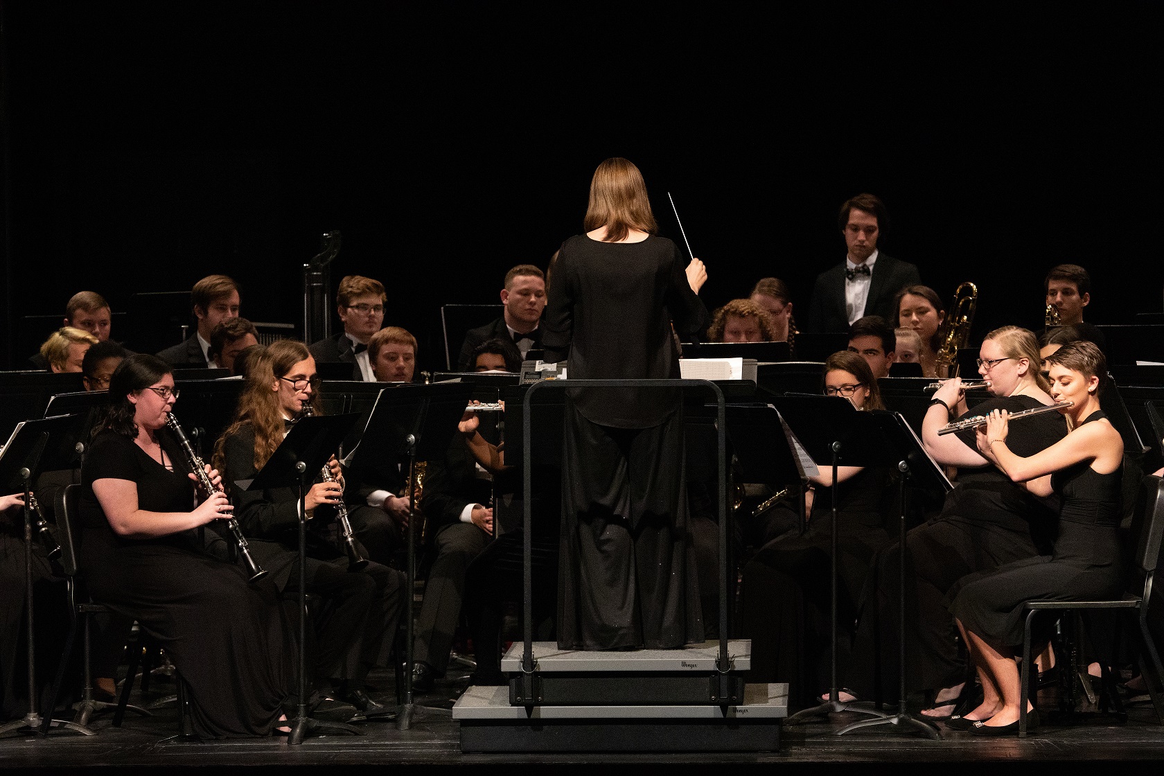 A band director is center stage with her back to the audience. Band members playing various woodwind instruments face her. They are all dressed in black