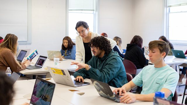 Professor helping a student while looking at a laptop surrounded by other student in the classroom
