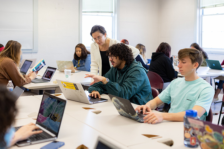 Professor helping a student while looking at a laptop surrounded by other student in the classroom