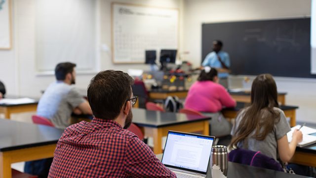 Student with laptop in the foreground with teacher and chalkboard in the background