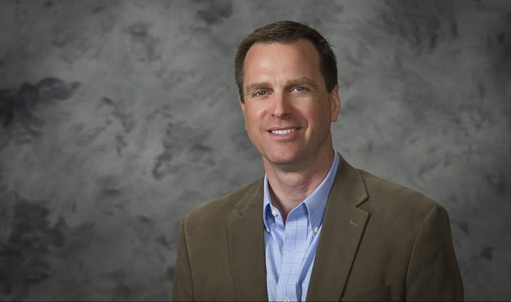 A headshot of Jamie Frueh. He is smiling while wearing a blue button up shirt and grey jacket