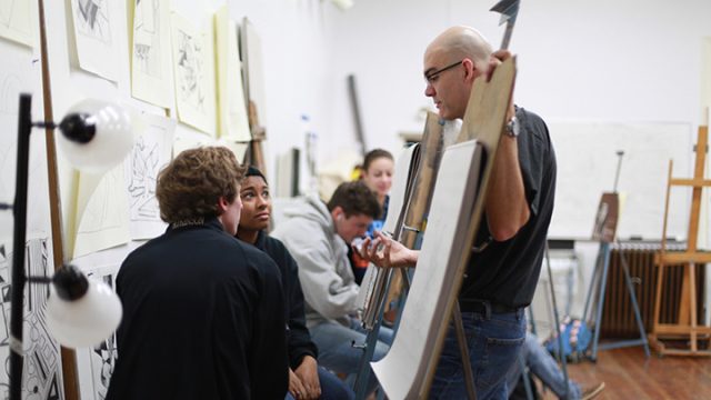 Professor speaks with students in art class standing behind an easel with paper on it