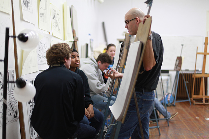 Professor speaks with students in art class standing behind an easel with paper on it
