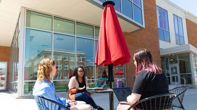 Three students sitting at a table outside of a building with glass windows