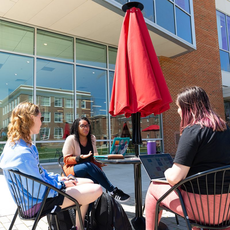 Three students sitting at a table outside of a building with glass windows