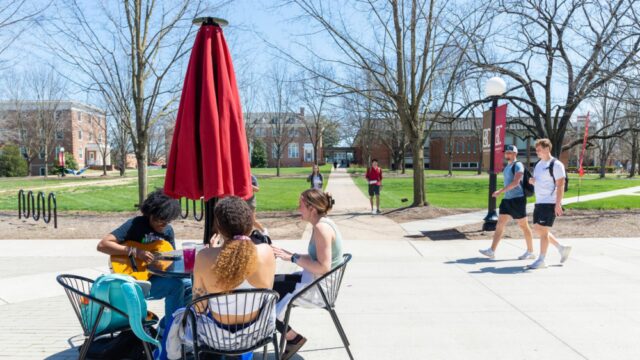 Three students sitting around a umbrella table with more students walking in the background