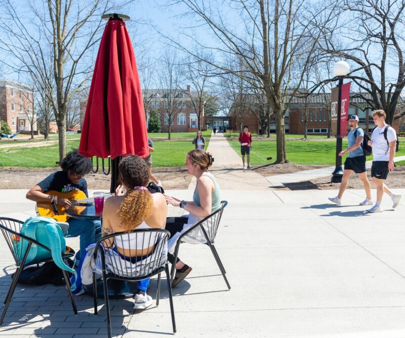 Three students sitting around a umbrella table with more students walking in the background