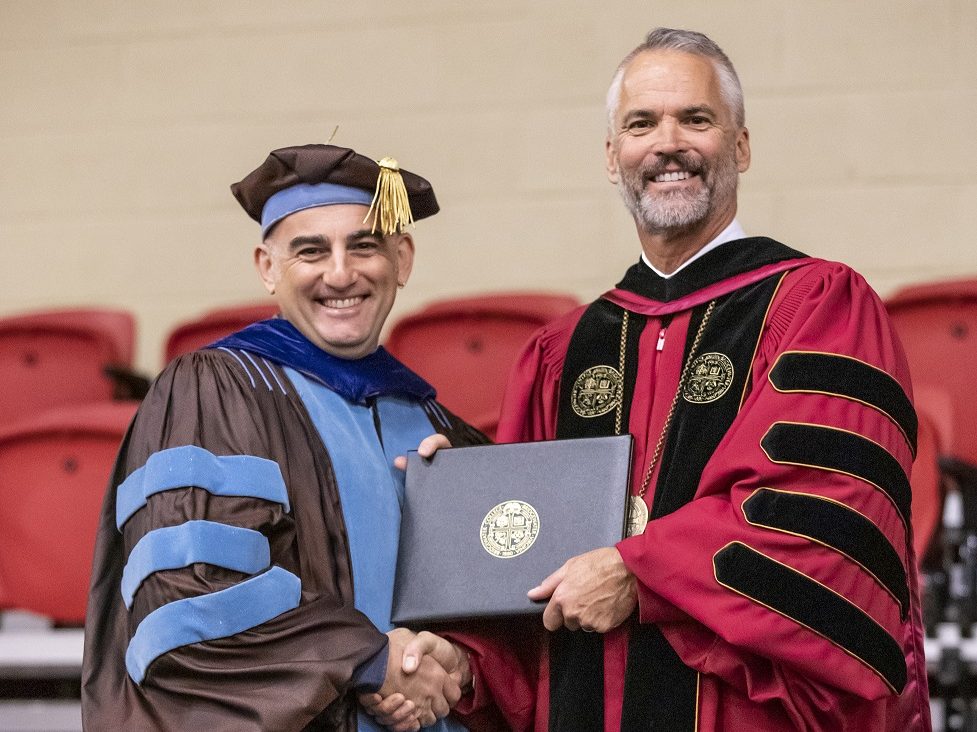 Two men shaking hands while accepting an award. Both are wearing academic regalia