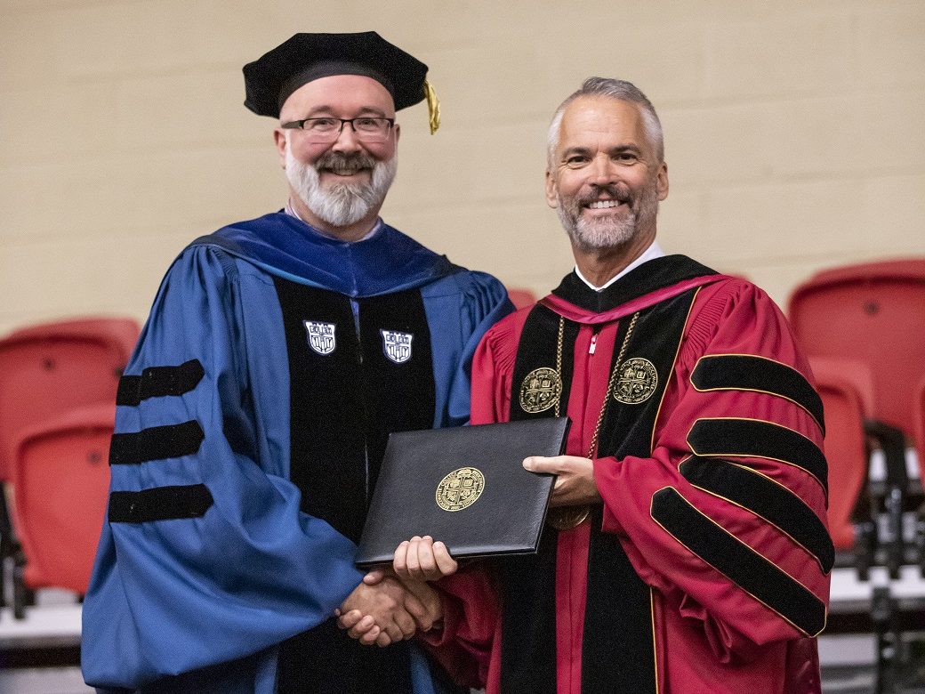 Two men shaking hands while accepting an award. Both are wearing academic regalia