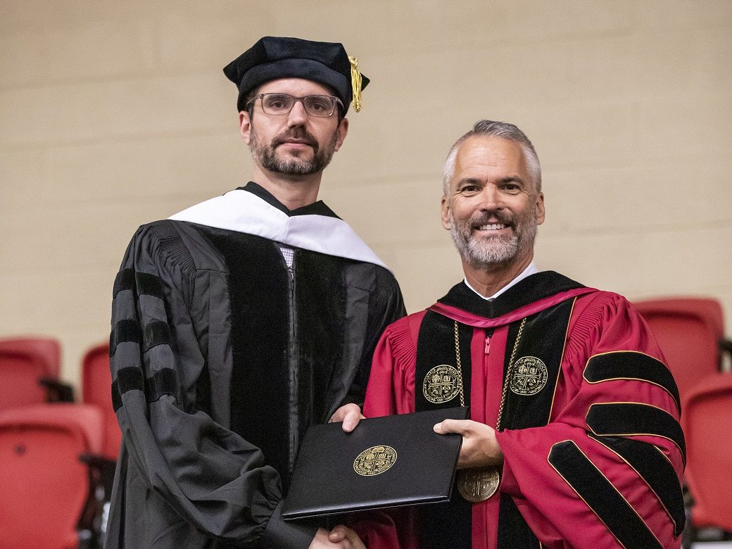 Two men shaking hands while accepting an award. Both are wearing academic regalia