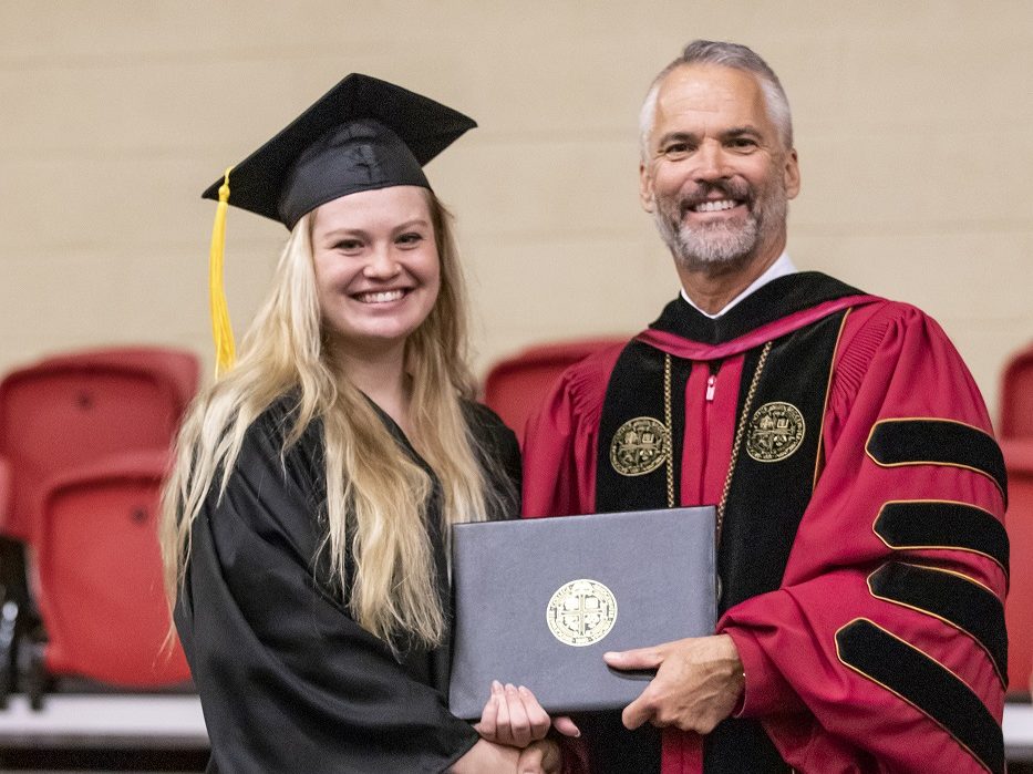 A women on the left and a man on the right shaking hands while accepting an award. Both are wearing academic regalia