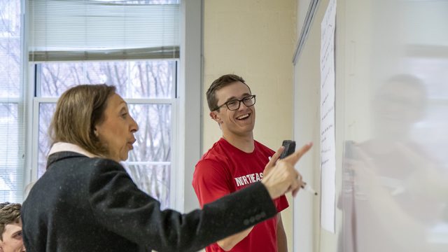 Professor in the foreground pointing to a white board with student smiling in the background