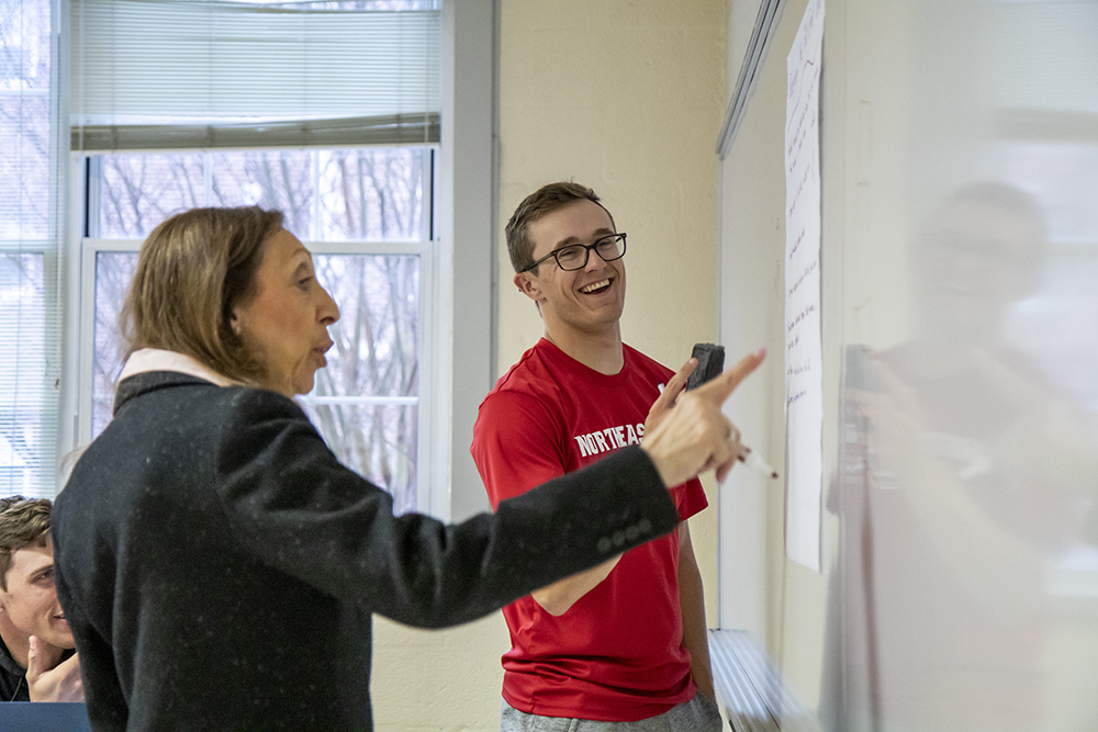 Professor in the foreground pointing to a white board with student smiling in the background