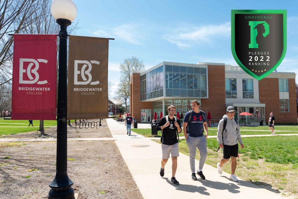 3 male students walk on campus. flags that say B-C are visible with the library in the background