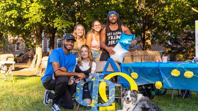 Group of students posing for photo with a sign and dog in front