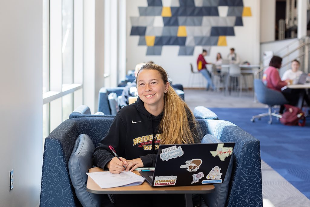 Female student working on laptop, taking notes, and smiling for the camera