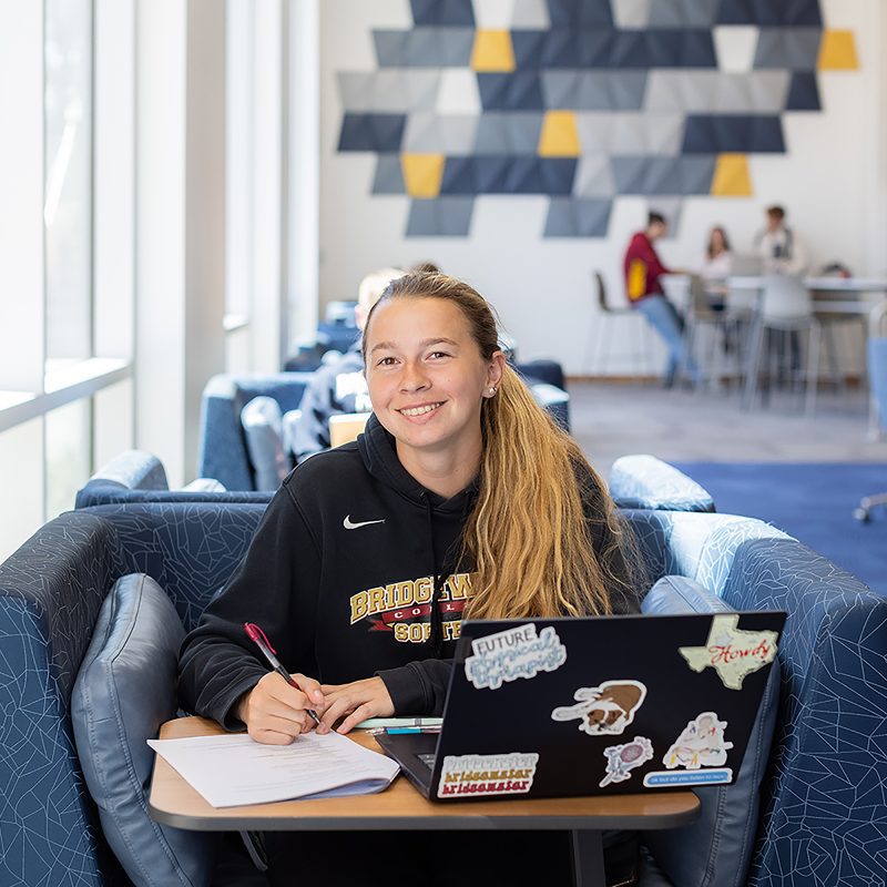 Female student working on laptop, taking notes, and smiling for the camera