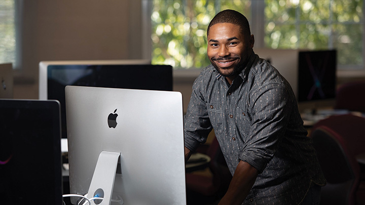 Man standing and smiling in front of Mac desktop computer