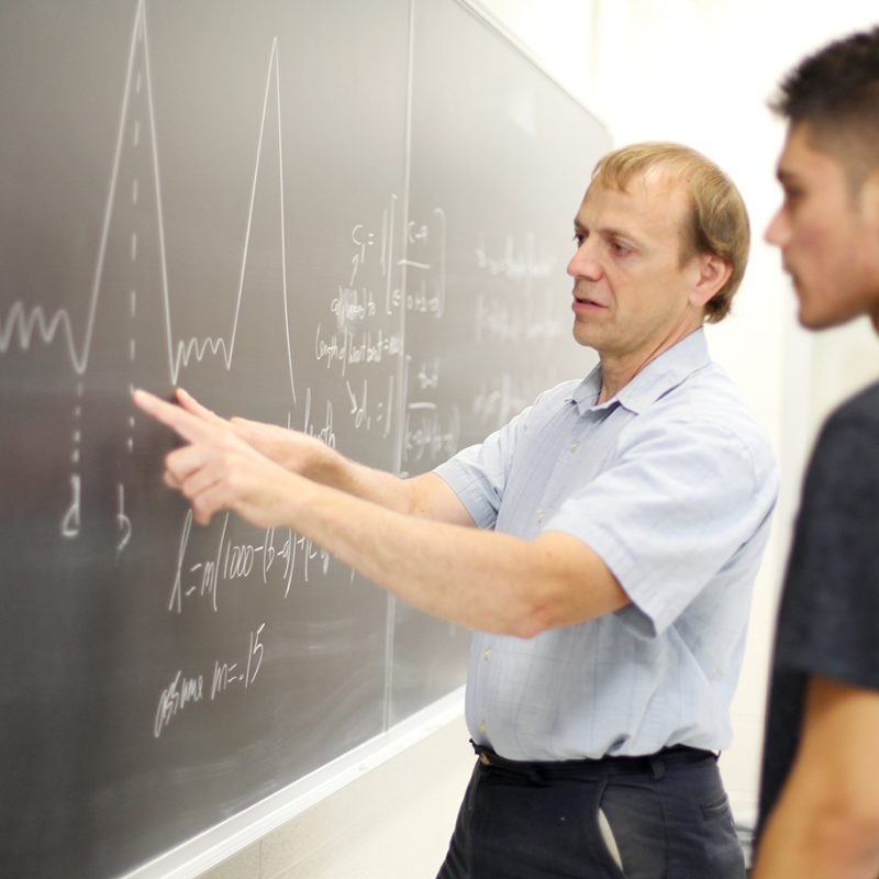 Professor pointing to chalkboard with math equations on it with student looking