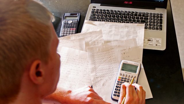 Student typing on calculator with papers and computer on the table