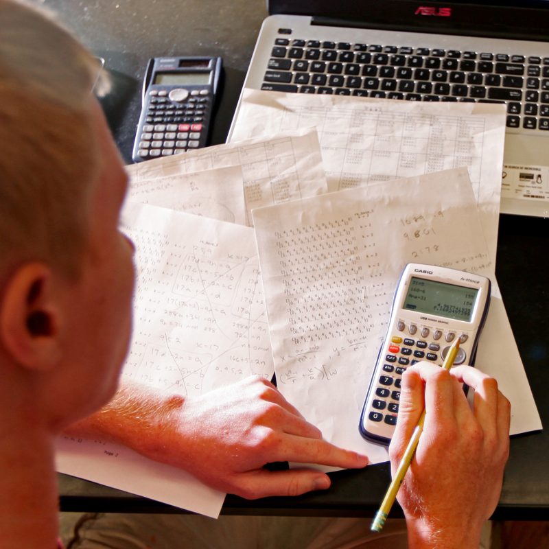 Student typing on calculator with papers and computer on the table