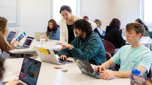 Professor helping a student while looking at a laptop surrounded by other student in the classroom